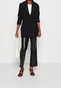 Cardigan nero lavorato a maglia con colletto a scialle, vita con cintura e due tasche frontali. Abbinato a pantaloni in ecopelle neri e sandali con punta aperta.