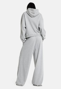 Άτομο με μακριά σκουρόχρωμα μαλλιά, ντυμένο με χαλαρό γκρι φούτερ με κουκούλα και φαρδιά γκρι φόρμα, στέκεται γυρισμένο προς τα πίσω σε λευκό φόντο.