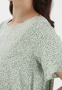 Haut vert clair avec un col rond et des manches courtes, orné d'un motif texturé de petits points blancs.