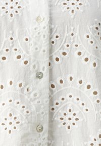 Chemise blanche brodée avec des motifs floraux complexes, texture douce et petites boutons de fermeture de couleurs contrastées.