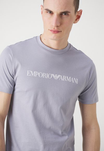 Heledane lilla puuvillane t-särk, millel on rinnal valge "EMPORIO ARMANI" logo, klassikaline ümar kaelus ja lühikesed varrukad.