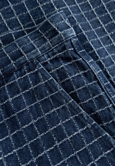 Dark blue denim fabric with white grid stitching, showing overlapping sections and seam details.