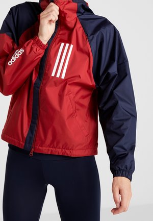 Osoba nosząca czerwoną i granatową kurtkę Adidas z białymi paskami, zapinając kurtkę, w granatowych legginsach.