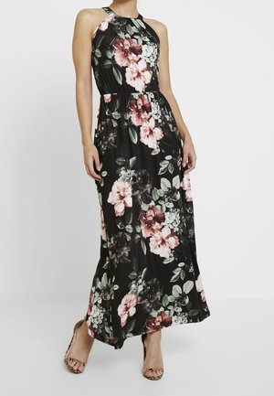 Vrouw draagt een zwarte mouwloze halternek maxi-jurk met roze, witte en groene bloemenprint, gecombineerd met open-toe hakken.