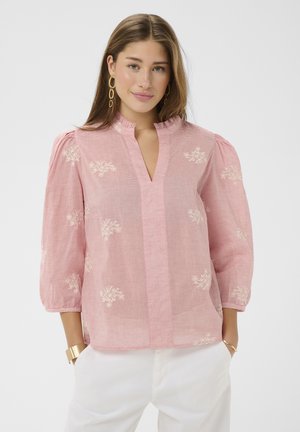 Femme portant un chemisier rose à manches trois-quarts bouffantes, avec une broderie florale subtile, un pantalon blanc, des boucles d'oreilles en or et des bracelets, sur un fond uni.