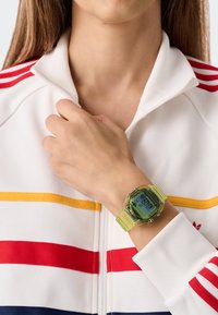 Ceas de mână digital cu carcasă și curea din plastic verde translucid. Dispune de un afișaj digital și este purtat pe o mână, lângă o jachetă albă cu dungi.