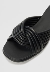 Sandale noire à semelle plate avec des brides en cuir souple entrelacées, un design à orteil arrondi et une texture lisse.