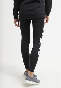 Svart leggings med en slät textur, med vit diagonalt tryck på sidan, matchat med grå sneakers för en avslappnad stil.