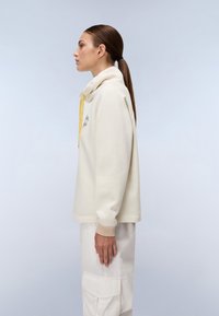 Kremowy sweatshirt z wysokim kołnierzem, żółtym sznurkiem ściągającym i ściągaczami. Noszony z jasnymi spodniami cargo, o minimalistycznym designie.