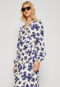 Robe enveloppante à manches longues en tissu crème, avec de grands motifs floraux bleus et noirs, dotée d'un détail noué à la taille et d'une texture douce.