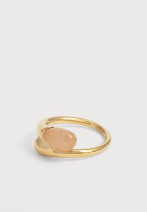 Bague de couleur or avec une pierre précieuse pêche lisse en forme ovale, sertie dans un design de bande ouverte sur un fond uni.