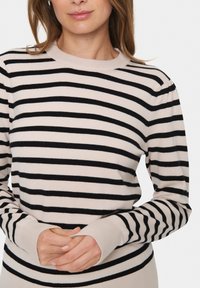 Hellbeige Pullover mit schwarzen Streifen, langen Ärmeln und gerippten Bündchen. Weicher Strick mit Rundhalsausschnitt. Hände auf der Taille verschränkt.