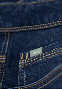 Närbild av en mörkblå denim-jeansficka med orange sömnad och en grå tagg märkt "Garcia" fäst vid fickans sömnad.