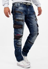 Pantalons cargo en denim bleu avec de multiples poches zippées, des accents en tissu noir renforcé et un design de poignet ajusté. Matériau texturé et durable.
