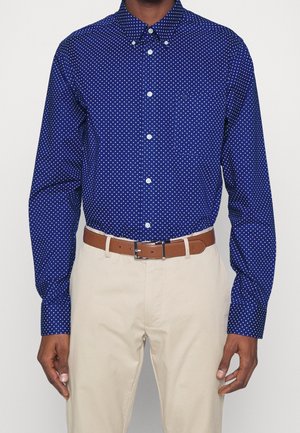 Mężczyzna ubrany w niebieską koszulę z długim rękawem zapinaną na guziki z małymi białymi wzorami trójkątów, beżowe spodnie i brązowy skórzany pasek.