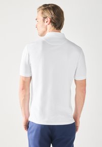 Camisa polo blanca de manga corta con un cuello moderno, textura suave y corte recto, mostrada desde la parte de atrás.