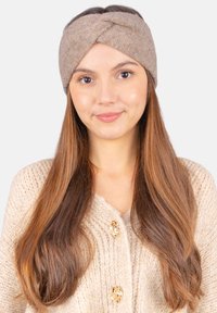 Bandeau en tricot beige avec un design torsadé. Fabriqué à partir d'un matériau doux, présentant une texture lisse. Porté avec un cardigan de couleur crème.