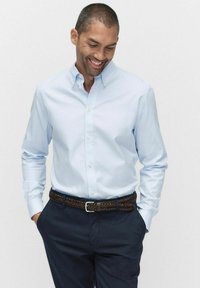 Lichtblauw overhemddes, lange mouwen, getailleerde pasvorm. Draagt een gevlochten bruine riem en donkerblauwe broek. Gladde stoftextuur.