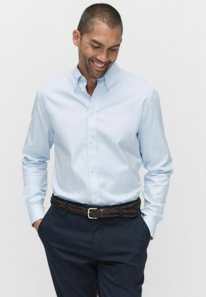 Chemise bleu clair à manches longues, coupe ajustée. Portée avec une ceinture tressée marron et un pantalon marine foncé. Texture de tissu lisse.