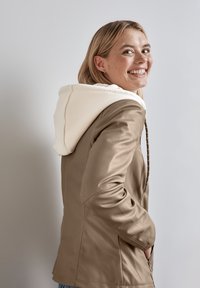 Giacca color marrone chiaro con cappuccio bianco, caratterizzata da una texture morbida e dettagli con zip. Presenta una vestibilità casual e tasche laterali.