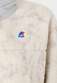Fleece pullover in light beige with a textured surface. Features a gray collar and a contrasting blue, red, and white logo on the chest.