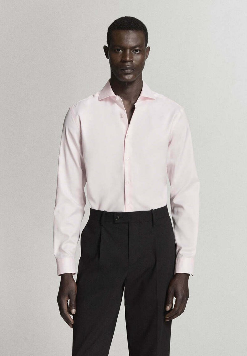 Homme portant une chemise rose clair à manches longues boutonnée, rentrée dans un pantalon noir taille haute, debout devant un fond uni.