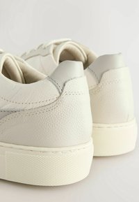 Baskets en cuir blanc avec finition texturée, accents gris et semelle en caoutchouc épaisse à motif. Détails cousus au niveau du talon.