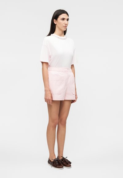 Γυναίκα στέκεται φορώντας ένα λευκό τοπ με κοντά μανίκια και λεπτομέρειες με κολιέ, ανοιχτό ροζ σορτς με τσέπες και σκούρα καφέ loafers με φούντες.