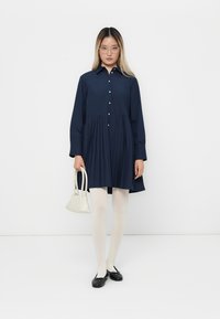Woman with long hair wearing glasses, navy pleated dress, white tights, black flats, holding small white handbag against plain white background.