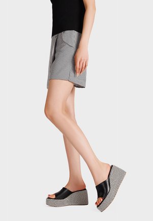 Donna che indossa un top nero, pantaloncini a quadretti bianco e nero e sandali con zeppa neri con tacchi a piattaforma decorati, su sfondo bianco.