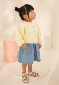 Gul strikket cardigan med puffede ærmer, kombineret med et denimskørt med blomsterbroderi og knappedetaljer. Multifarvede sandaler med stropper.