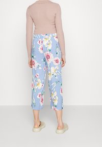 I pantaloni floreali presentano uno sfondo azzurro chiaro con grandi fiori rosa e gialli, vestibilità rilassata e lunghezza fino al polpaccio, indossati con zoccoli beige.