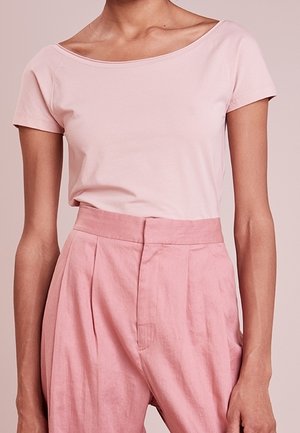 Kvinde iført en lyserød kortærmet top og løse, højtaljede pink bukser, stående mod en ensfarvet baggrund.