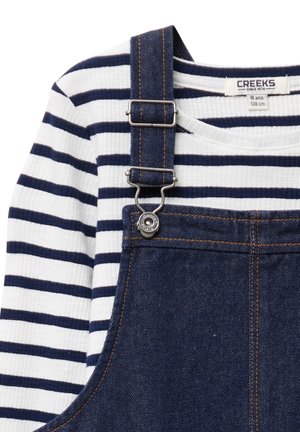 Marineblauwe denim salopettes met verstelbare banden, zilverkleurige hardware en een witte langemouwen shirt met marineblauwe horizontale strepen.