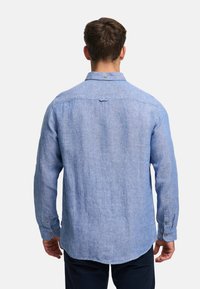 Mies nähtynä takaa, hänellä on päällään vaaleansininen pitkähihainen pellavapaita ja tummat housut yksinkertaista vaaleaa taustaa vasten.