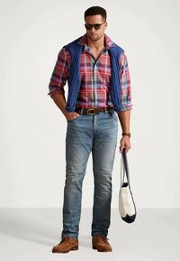 Moški nosi sončna očala, rdečo karirasto srajco z modrim puloverjem, ki ga ima ovitega okoli ramen, jeans hlače, rjave čevlje in drži belo-modro platneno torbo.