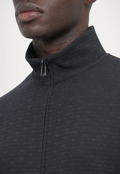 Giacca nera con zip e collo alto, caratterizzata da un motivo logo ripetuto, tessuto strutturato e una vistosa cerniera argento.