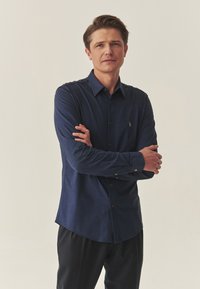 Tumši zils garām piedurknēm krekls ar pogājamu apkakli un mazu izšūtu logotipu uz krūtīm, kombinēts ar tumšiem biksēm.