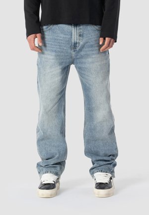 Homme portant un jean bleu clair ample, une chemise à manches longues noire et des baskets noir et blanc, debout devant un fond uni.