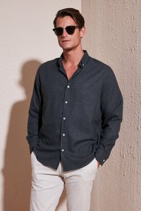 Camisa abotonada de color gris oscuro con cuello, mangas largas y botones blancos, combinada con pantalones de tonos claros y gafas de sol negras.