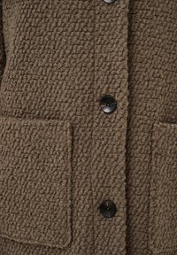 Manteau en laine marron texturé avec une grande poche avant et deux boutons noirs fermés au centre.