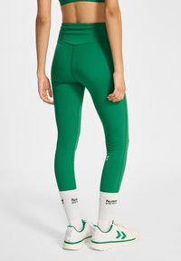 Zielone legginsy z wysokim stanem, gładką fakturą i dopasowanym krojem. Połączone z białymi skarpetkami i trampkami z zielonymi wykończeniami.