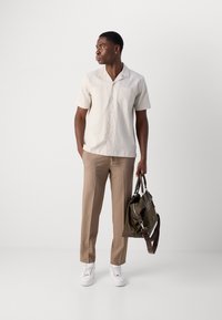 Camisa de manga corta texturizada beige, pantalones cortos marrones a medida, zapatillas blancas y un bolso verde oliva con bandolera.