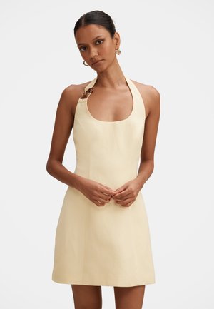 Kvinde iført en ærmeløs beige halterkjole med en dekorativ spænde på den ene strop, stående med hænderne løst foldet foran sig.