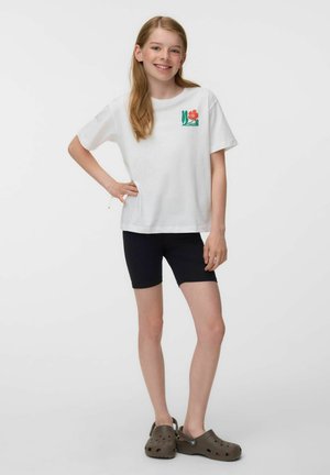 Meisje met lang blond haar staat, draagt een wit T-shirt met bloemmotief, zwarte shorts en bruine instapschoenen.