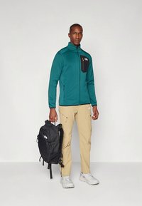 Teal fleece jacket with black zip pocket, beige pants, and black backpack. Model wears light grey sneakers. Simple design, no patterns.