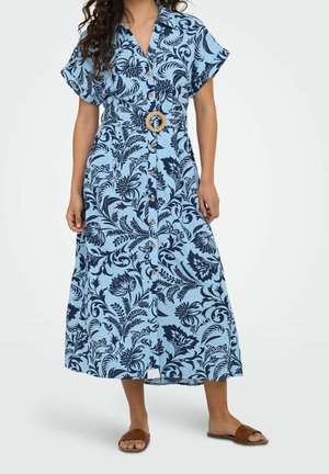Femme portant une robe midi bleu clair à imprimé floral bleu marine, avec boutonnage devant, manches courtes et une ceinture circulaire tissée, assortie à des sandales marron.