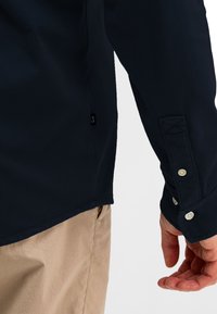 Man draagt een marineblauw lang mouw shirt met een gebuttonde manchetten en beige broek, hand ontspannen langs zijn zij tegen een neutrale achtergrond.