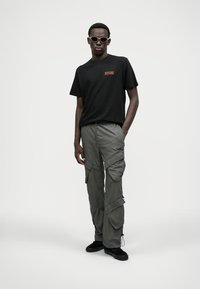 T-shirt en coton noir avec un logo rouge, associé à un pantalon cargo olive avec poches et poignets réglables. Des baskets noires complètent le look.