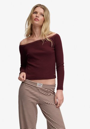 Lyshåret kvinde iført en burgundy off-shoulder langærmet top og brune og beige stribede bukser, stående mod en hvid baggrund.